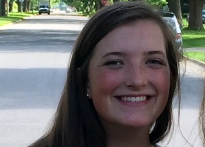 A warm, professional photo of professional. She has long brown hair, and is smiling directly at the camera against a greenery backdrop