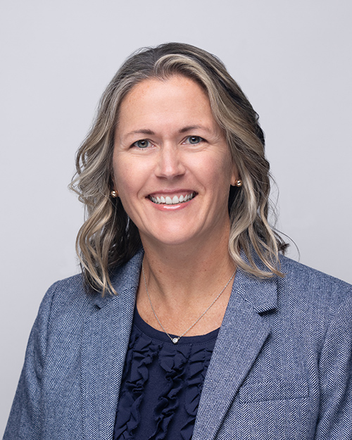 Professional headshot of Jennifer Goldbach. She is smiling with shoulder-length wavy hair and is wearing a blue textured blazer over a navy ruffled blouse with a simple necklace, against a light grey background.