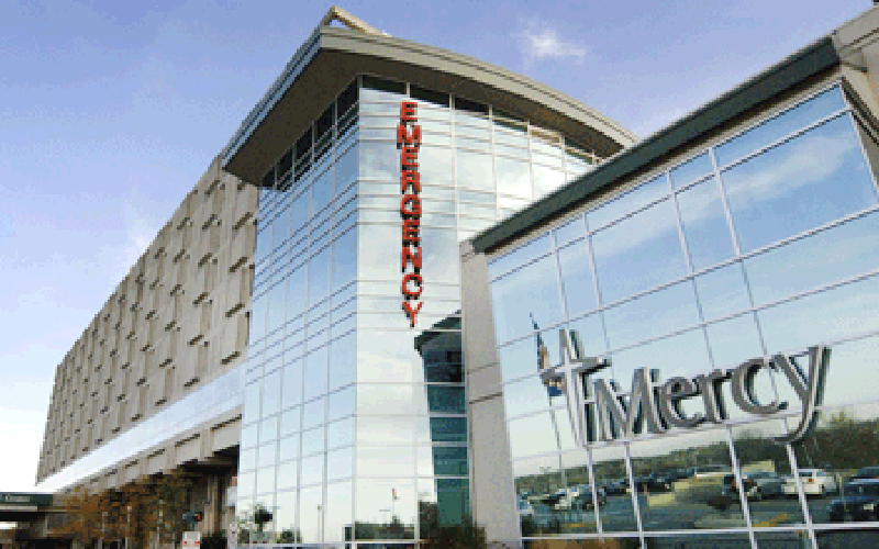 Modern hospital building with large glass facade, reflecting a clear sky. Bold red 