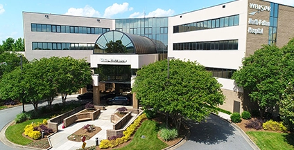 A modern office building with large glass windows and a curved atrium above the main entrance. Lush green trees and manicured gardens surround the entrance.