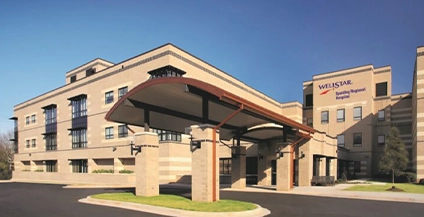 Hospital entrance with a modern structure, featuring a curved canopy supported by pillars. The building facade is beige brick, set against a clear blue sky.