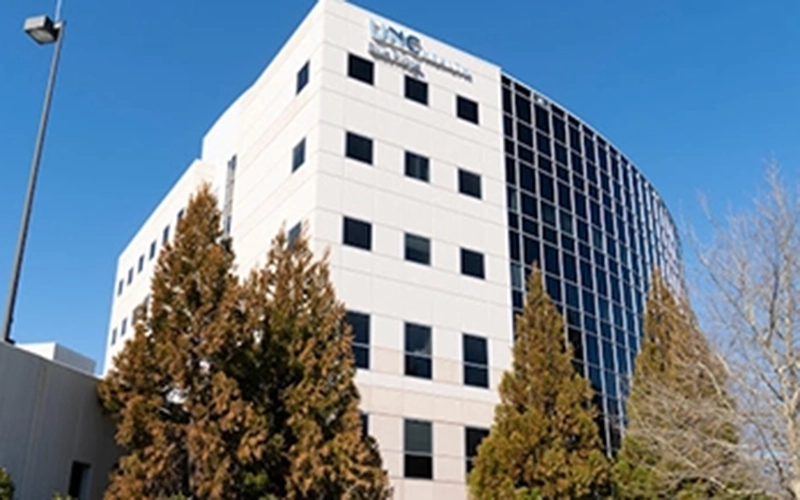 A modern, multi-story hospital building with a mix of white walls and dark glass windows. Foreground includes tall, leafy trees. Bright blue sky.