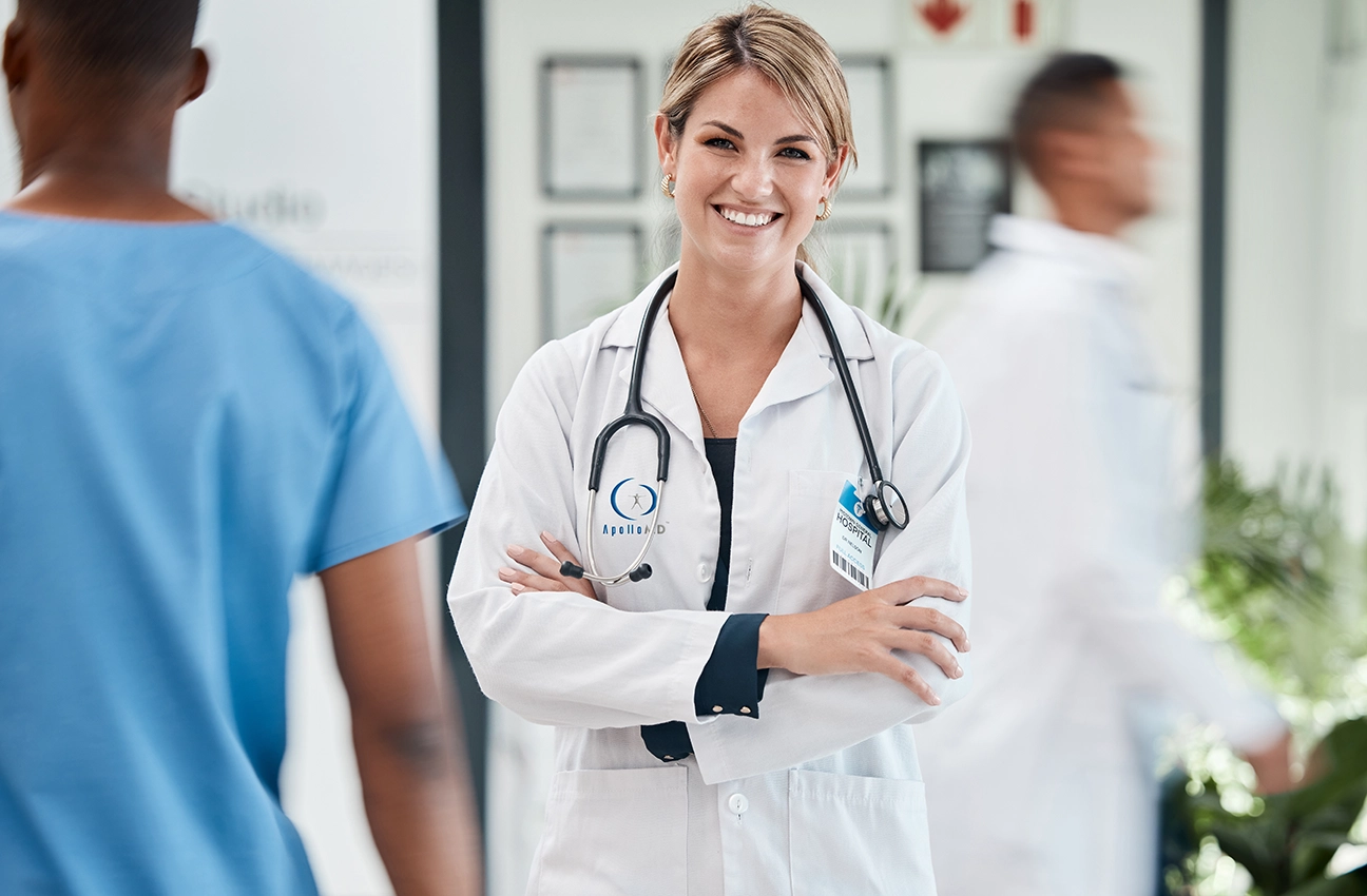 Smiling female doctor with a stethoscope stands confidently in a hospital hallway, arms crossed. Blurred movement of hospital staff conveys busy atmosphere.