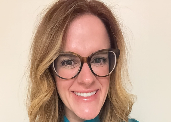 A woman with long, wavy light brown hair and glasses smiles at the camera. She is wearing a teal top, set against a plain, light-colored background.