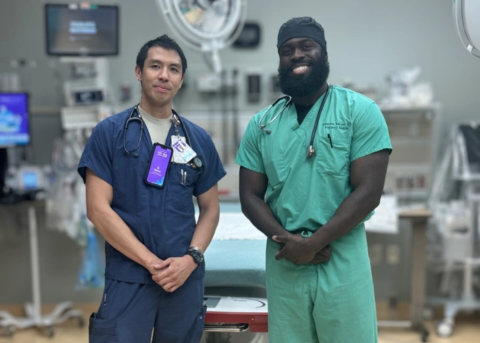 Two medical professionals, one in navy scrubs and the other in green, stand smiling in a hospital room with medical equipment in the background.