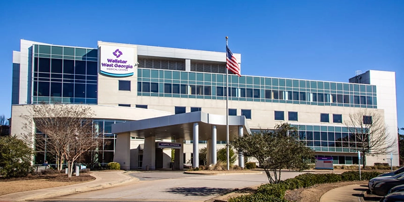 Modern hospital building with glass and concrete facade under clear blue sky. An American flag stands in front, with paths and trimmed shrubs nearby.
