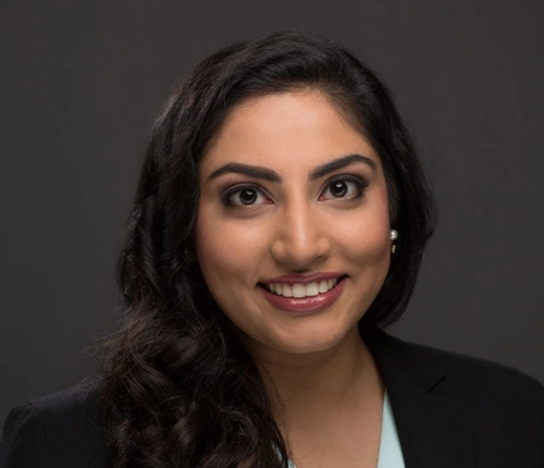 Amna Jamshad, Smiling woman with long, wavy dark hair, wearing a black blazer and light top, against a dark background. She conveys confidence and warmth.