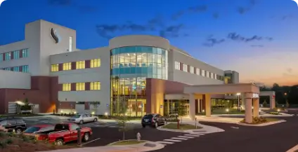 A modern hospital building at dusk, with large glass windows illuminated from within. Several cars are parked in the foreground under a twilight sky.