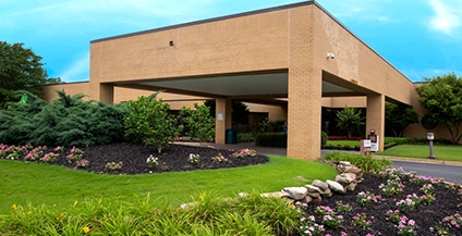 A low, modern brick building with an open carport is surrounded by well-kept gardens, including vibrant flowers and lush green shrubs. Clear blue sky overhead.