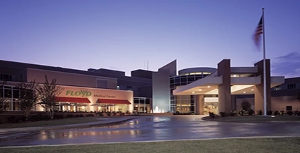 Exterior of Floyd Medical Center at dusk. The building is lit with soft lights, an American flag waves on the right, and the sky is a gradient of blue and purple.