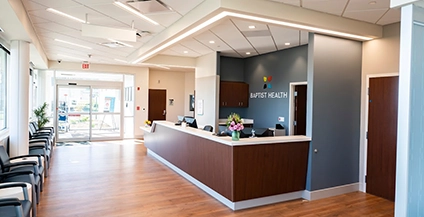 A modern, bright hospital lobby with a wooden floor, a central reception desk adorned with flowers, chairs lined up on the left, and large windows offering natural light.