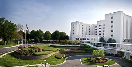A modern hospital building with a white facade, surrounded by landscaped gardens and a curved driveway. Clear sky and trees in the background.