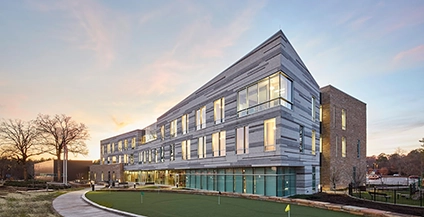 Modern, angular building with large windows at sunset, surrounded by a manicured lawn. The sky is colorful, creating a calm, serene atmosphere.
