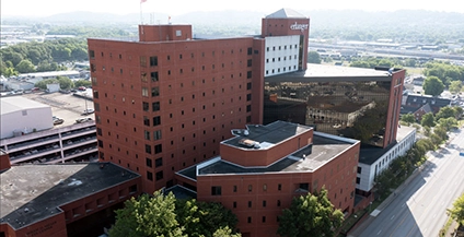 Aerial view of a large red-brick building complex with trees in the foreground. A digital facade display shows a historical scene. Bright, clear day.