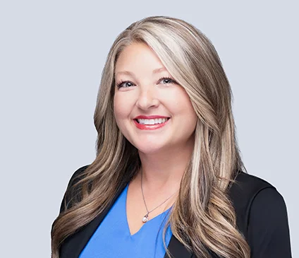 A professional headshot of Jamie Wise. She is smiling with long, blonde hair and is wearing a bright blue top with a black blazer and a pearl necklace against a light grey background.