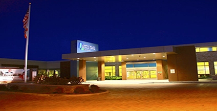 A modern building entrance at night, warmly lit with yellow lights. An American flag is on the left, and the sky is a dark blue, creating a welcoming atmosphere.