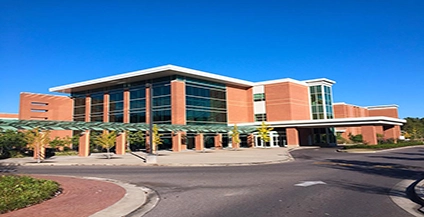 Modern brick and glass building under a clear blue sky, featuring a covered walkway, surrounded by landscaped greenery and an empty paved driveway.