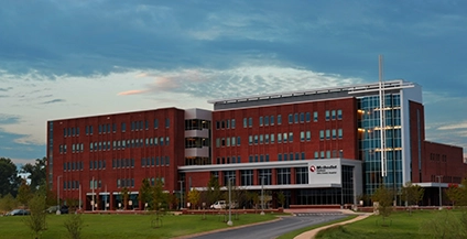 A modern red-brick hospital building with large windows stands under a cloudy sky, surrounded by greenery. The scene conveys a sense of calm and professionalism.