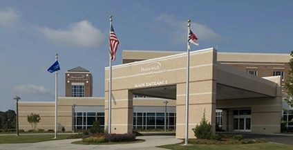 Wide entrance of a modern hospital building with a large canopy, flanked by three flagpoles. Clear sky overhead, conveying a welcoming atmosphere.