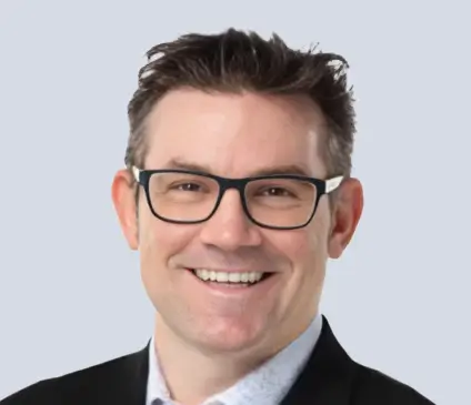 A bright, approachable portrait of Aaron Warmus. He has a wide, warm smile and brown hair, wearing black rimmed glasses and a black blazer. He is looking directly at the camera against a neutral grey studio backdrop.