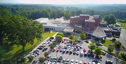 Aerial view of a large red-brick school with a busy parking lot and lush green trees around. A bright, clear sky suggests a sunny day.