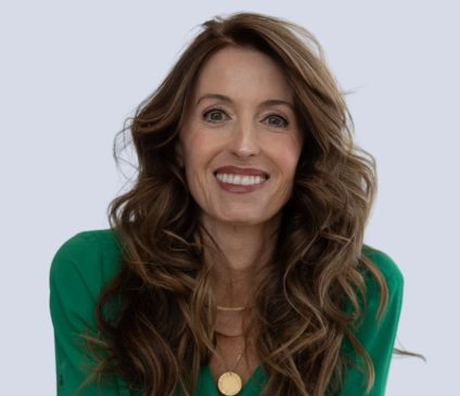Stefaney Caro, A woman with long, wavy brown hair and wearing a green blouse smiles warmly at the camera. She has a gold necklace and a calm, friendly expression.