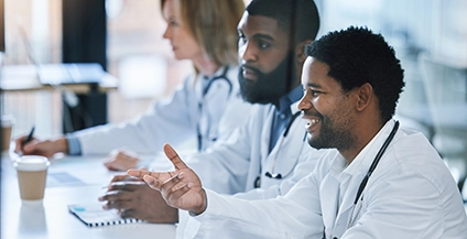 Three doctors in white coats sit at a conference table, engaged in discussion. The setting is bright and professional, conveying collaboration.