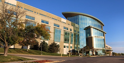 Modern hospital building with large glass windows and a curved roof, set against a clear blue sky. Trees flank the entrance, suggesting a calm environment.