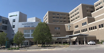 A large, multi-story hospital building with a spacious entrance, trees, and a clear blue sky. The design is modern, conveying a sense of care and professionalism.