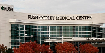 Front view of Rush Copley Medical Center, a modern building with large glass windows. Red autumn trees in the foreground, under a cloudy sky.