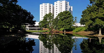 A modern white building with rounded edges stands behind lush trees, reflecting in a calm pond. Clear blue sky enhances the serene atmosphere.