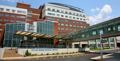 Large hospital building with a mix of red brick and white facade. Glass entrance and skywalk in front. Sign reads “Wilkes-Barre General Hospital.”