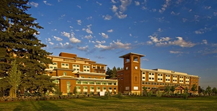 A large brick building with a clock tower stands on a grassy lawn under a blue sky with scattered clouds.