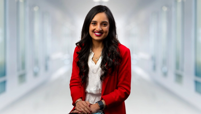 Harmisha Gandhi, PA-C, smiling in a red blazer and black top in a blurred hospital hallway.